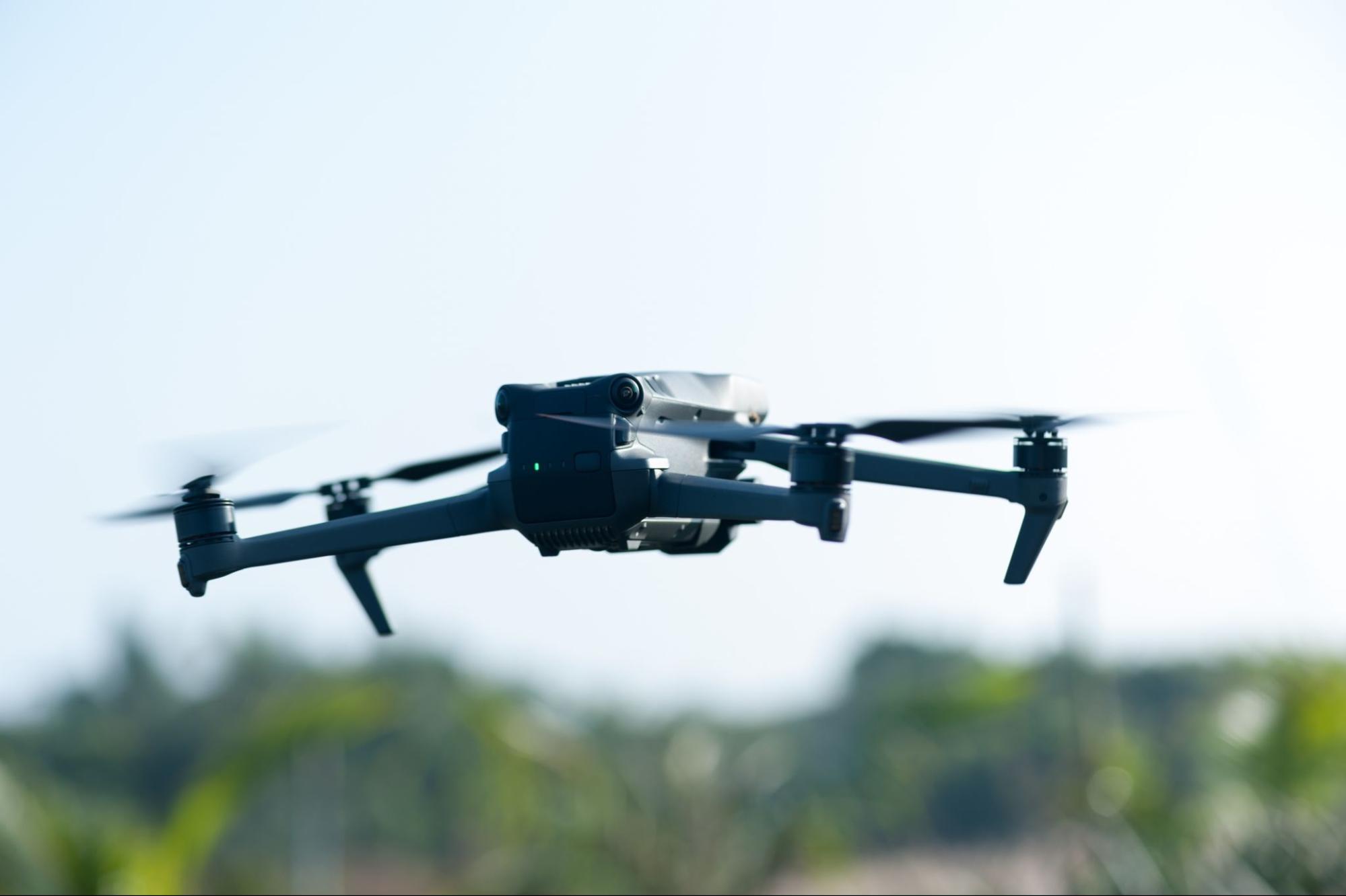 Een drone vliegt in de lucht met groene bomen op de achtergrond.