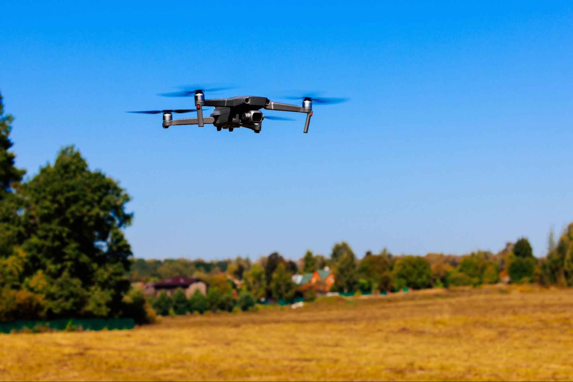 Een drone vliegt boven een open landschap met bomen en huizen op de achtergrond.
